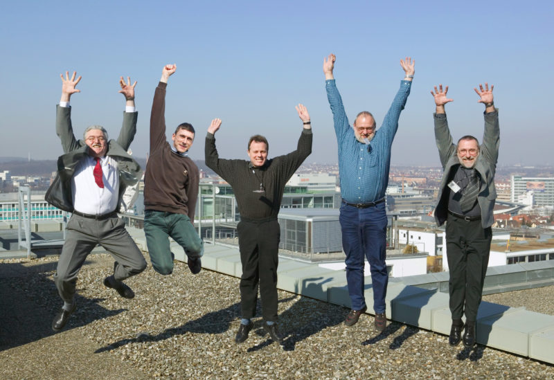 Gruppenfoto: 5 Mitarbeiter springen auf dem Dach ihres Betriebsgebäudes gleichzeitig in die Luft. Ihre umgehängten Betriebsausweise  fliegen umher.