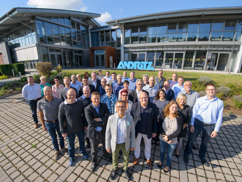 Group photo:  Staff in front of their company building. The boss stands in front, behind of all you can see the company building with large glass surfaces and the logo in front.