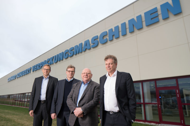 Group photo: The four managing directors of a medium-sized company outside in front of the company building on which the logo of the company is emblazoned.