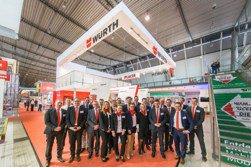 Group photo: Staff of an exhibition stand shortly before the opening of the exhibition to the public. In the background the entire exhibition stand.