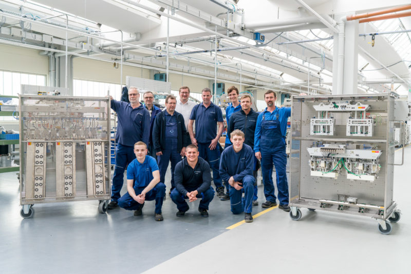 Group photo: Proud employees in the production of a company. They stand in the bright factory hall between rolling shelves with components. They all wear work clothes with the company logo sewn on them.