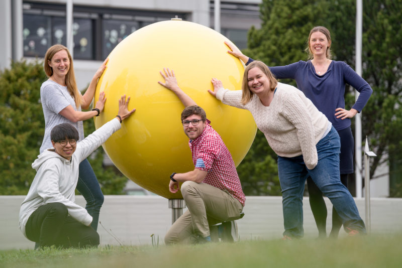 Group shot: Group photo: 5 employees of an astronomical research institute in front of the main building of the facility. They have their hands on a large yellow ball, a symbolic representation of the sun.