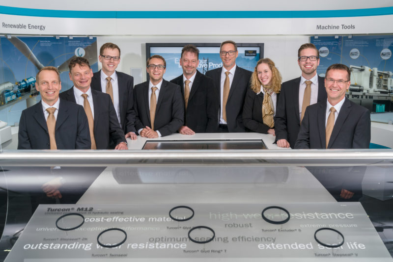 Group photo: Nine sales employees in the showroom of their company. In the preceding you can see products of the company. All wear official clothing with golden ties and scarves.