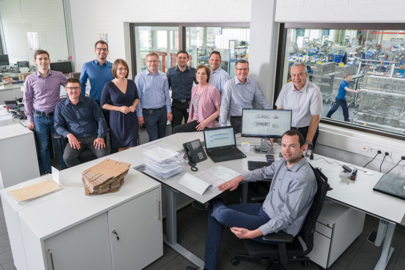 Group photo: Portrait of a group of 11 employees of a department. The picture was taken with a flash system, so that the people in the interior are still harmoniously illuminated and one can still look through the glass panes into the brightly lit production hall, in which another employee is moving a workpiece.