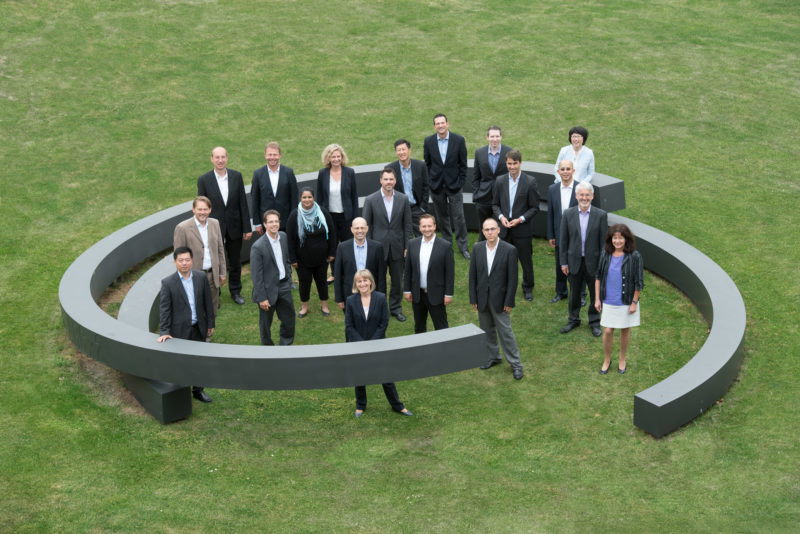 Gruppenfoto: Portrait von rund 30 Personen einer Firmenabteilung. Das Bild wurde mit langer Brennweite von einem in der Nähe stehenden Gebäude fotografiert. Die Personen stehen locker gruppiert innerhalb einer rund geformten großen künstlerischen Plastik auf dem grünen Rasen.