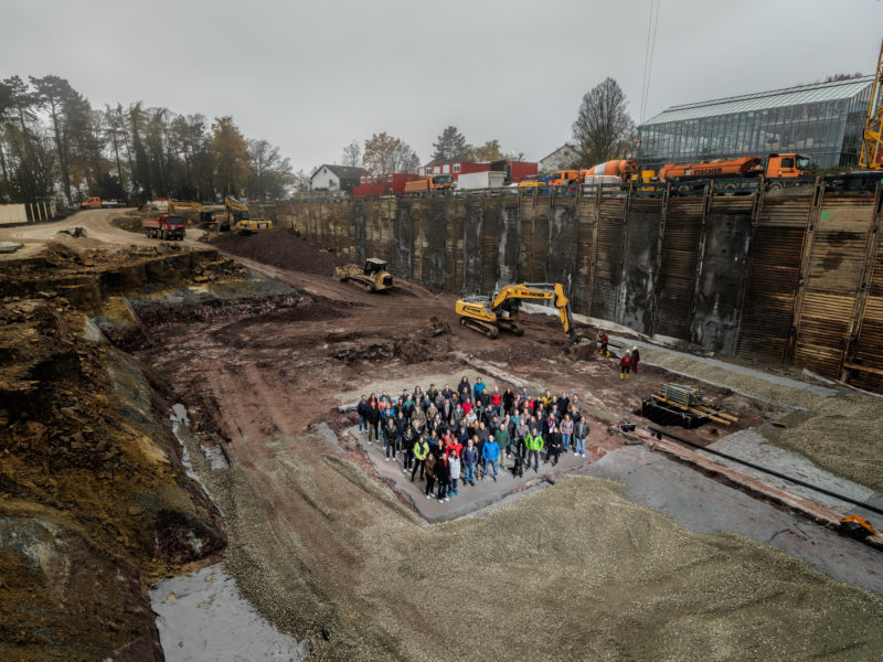 Gruppenfoto: Anlässlich des Baubeginns eines neuen Forschungsgebäudes posieren die zukünftig dort Beschäftigten für ein Gruppenbild auf der ersten Grundplatte des Gebäudekomplexes. Sie verlieren sich beinahe innerhalb der riesigen und tiefen Baugrube.