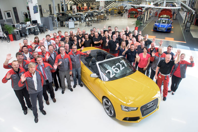 Group photo: The employees at the assembly line of an automibile production celebrate the construction of an anniversary vehicle. The car is in the middle and the colleagues wave into the camera, while you can see the production in the background.