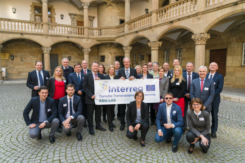 Gruppenfoto: Die Teilnehmer in der Pause einer internationalen Konferenz im Innenhof des historischen Schlosses, in dem sie tagen. Sie tragen ein Banner mit dem Namen der Veranstaltung.