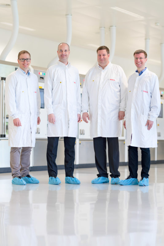 Gruppenfoto: Vier Mitarbeiter einer Abteilung, die an medizinischen Geräten arbeitet, stehen mit blauen Einmalüberziehern für die Schuhe und Schutzkitteln vor einer der Maschinen, die sie nutzen.
