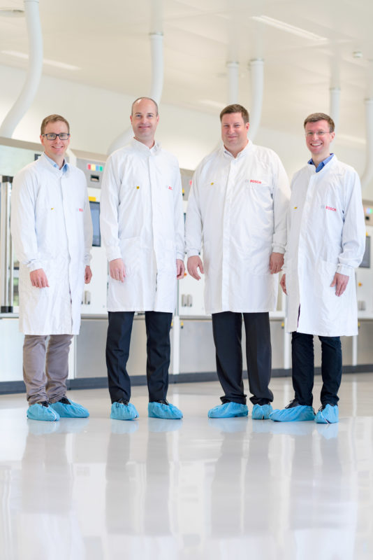 Group photo: Four employees in a department that works on medical equipment are standing in front of one of the machines they use with blue disposable covers for their shoes and protective coats.