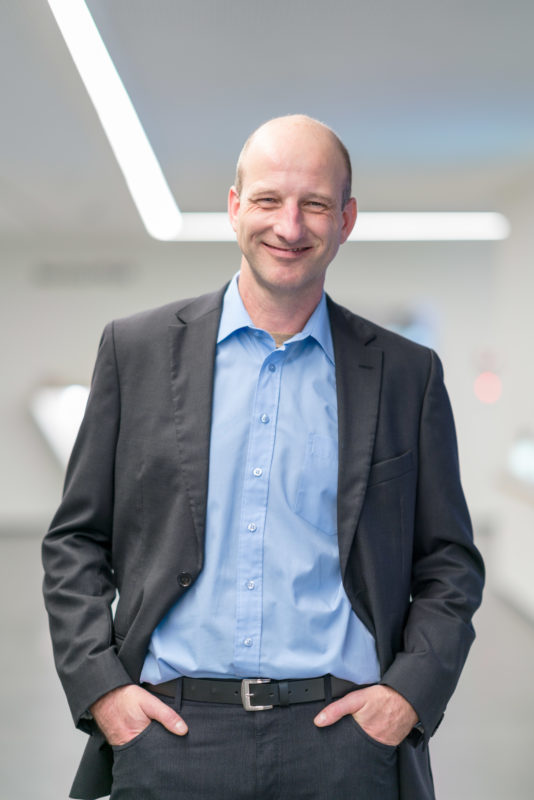 Managerportrait: A manager in half-portrait stands in the hallway of his modern and bright company building. His hands are casually in his pockets. The picture is taken with the fully open aperture of a fast telephoto lens and only slightly brightened with the flash.