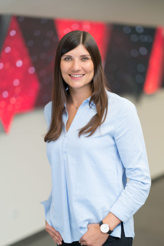 Portrait photography: Employee photography: Half portrait with fill-in flash of an employee standing in front of a glowing screen wall with the blurred content of a video in the foyer of a company. Recorded with existing lighting and additional studio flash system to improve the illumination of the person.