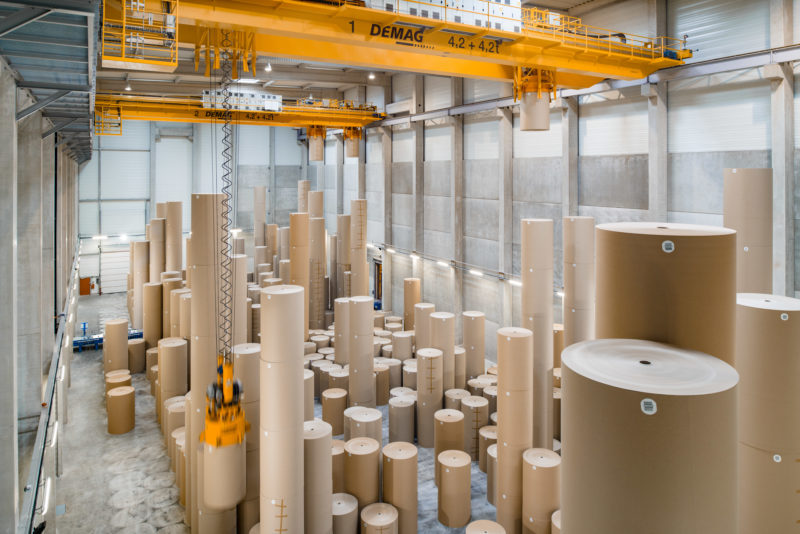 Industrial photography: Two indoor overhead traveling cranes move vertically stacked rolls of brown cardboard paper weighing several tons in a paper warehouse of a paper mill near Strasbourg.