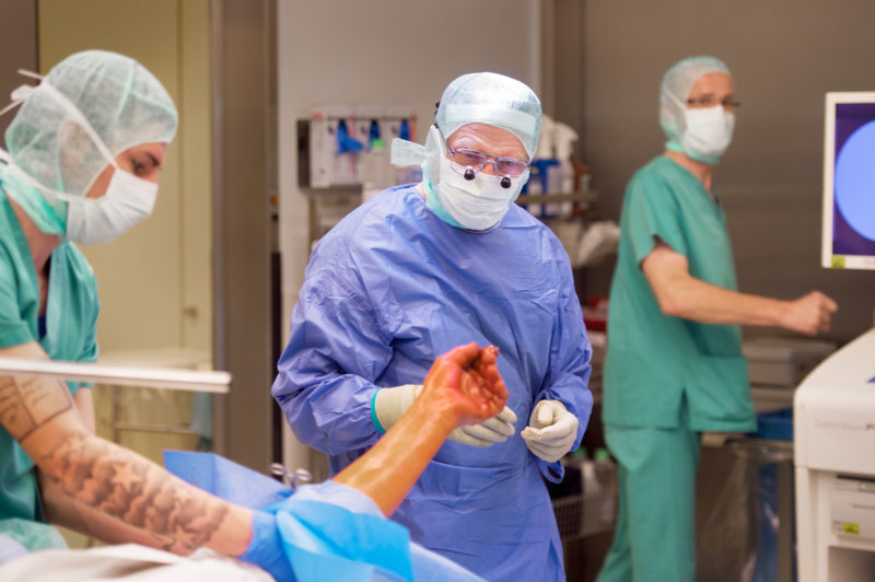 Medizinfotografie: Der Chirurg betrachtet vor dem Beginn der Operation die Finger der zu operierenden Hand. Er trägt eine Operationsbrille.