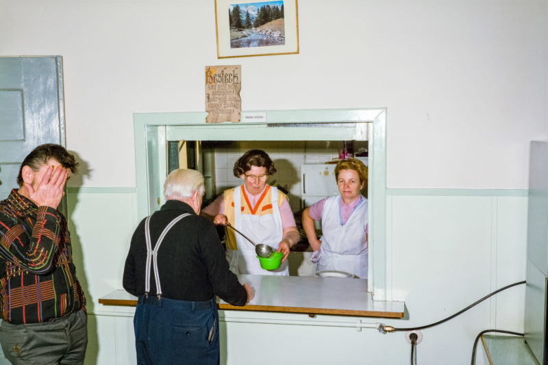 Editorial photography at the Salvation Army: Two women in white aprons serve soup to the needy. Above the window to the kitchen hangs a picture of a beautiful alpine landscape.