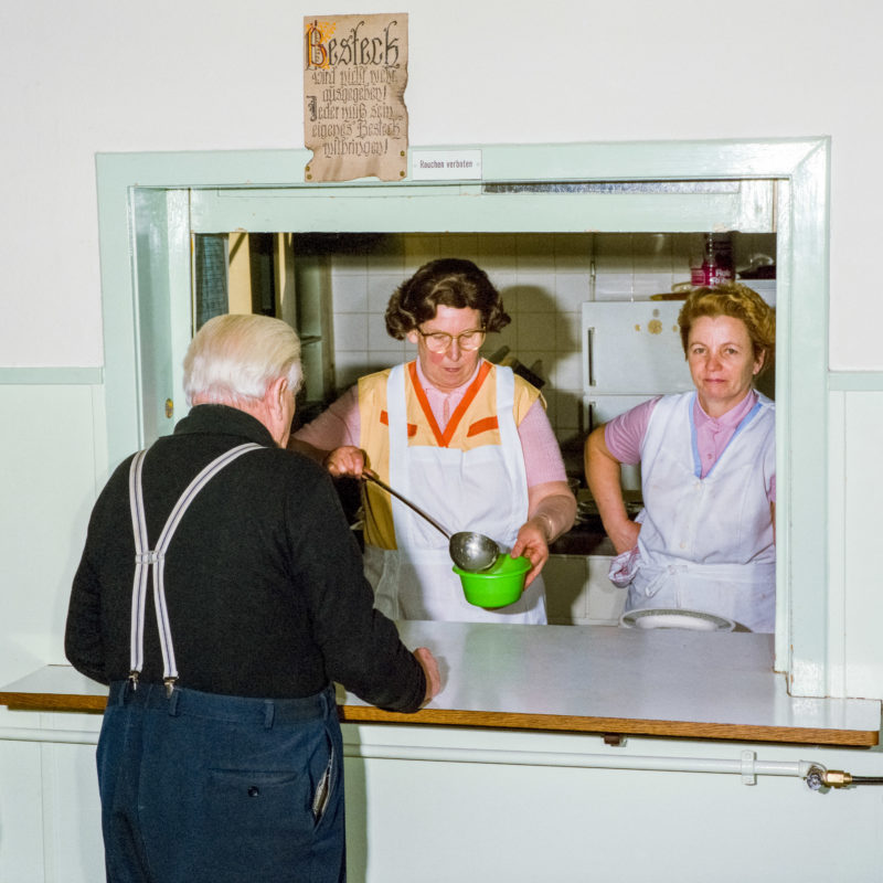 Luftaufnahmen und Drohnenfotografie: Reportagefotografie bei der Heilsarmee: Zwei Frauen in weißen Schürzen schenken Suppe an Bedürftige aus. Über dem Fenster zur Küche hängt ein Bild einer schönen Alpenlandschaft.