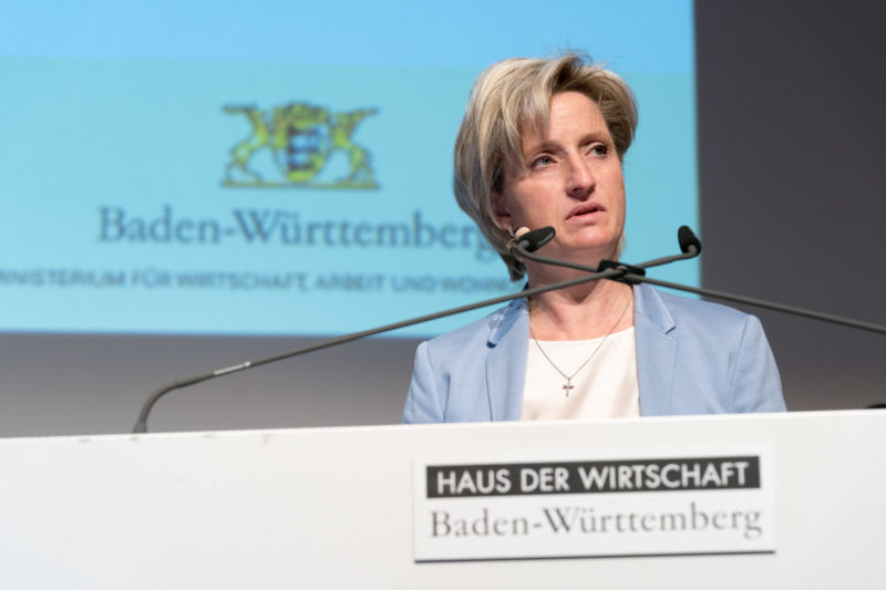 Eventfotografie, Veranstaltungsfotografie Reportagefotografie: Die Ministerin für Wirtschaft, Arbeit und Wohnungsbau des Landes Baden-Württemberg Dr. Nicole Hoffmeister-Kraut während einer Ansprache auf einer Veranstaltung im Haus der Wirtschaft in Stuttgart. Im Vordergrund sieht man das logo des Hauses und im Hintergrund das des Landes Baden-Württemberg.