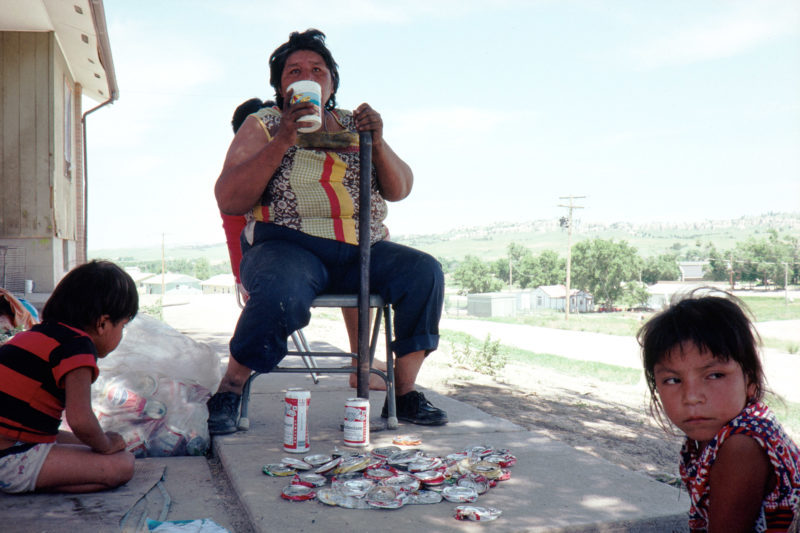 Reportage photography on slide film in the Pine Ridge Reservation in South Dakota, USA: A woman sits with children in front of the hut and crushes collected empty beer cans to earn some money by selling them.