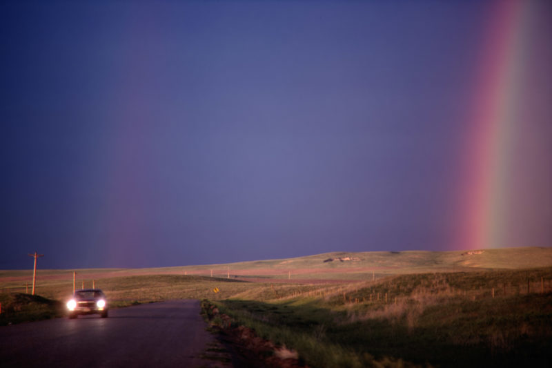Reportage photography on slide film in the Pine Ridge Reservation in South Dakota, USA: On one of the few tarred roads in the reserve, an old car drives with lights on, while the landscape behind it is covered by a rainbow.