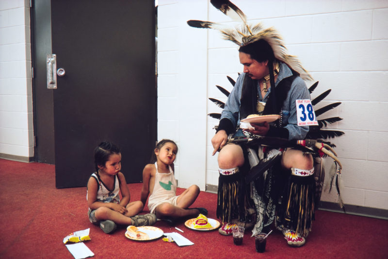 Reportage photography on slide film in the Pine Ridge Reservation in South Dakota, USA: During a dance competition a dancer in full ornament sits with his children in the corridor to eat from paper plates.