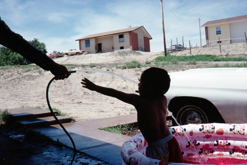 Reportage photography on slide film in the Pine Ridge Reservation in South Dakota, USA: A child plays with water from a hose while sitting for washing in a plastic paddling pool in a settlement.