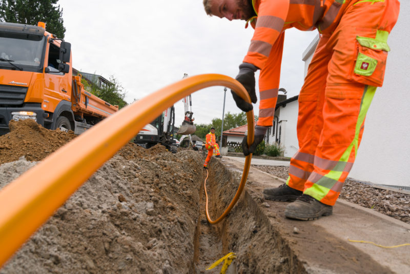 Industriefotografie: Für den Internet-Breibandausbau in ländlicher Regionen werden Glasfaserkabel unter einem Gehweg in einer Wohnsiedlung verlegt. Zwei Arbeiter senken ein Verlegerohr in den Graben, durch das später die Glasfaserkabel geblasen werden.