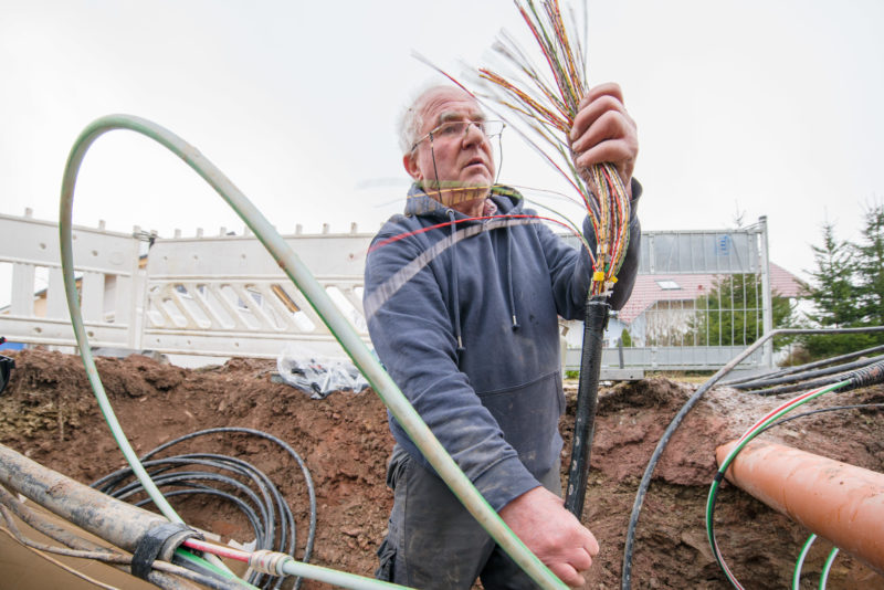 Industriefotografie: Internetausbau auf der Straße: Ein Techniker steht im ausgehobenen Graben und hat ein Kabelbündel mit Telefon und Datenleitungen in der Hand, um dieses an einem neuen Verteiler mit Glasfasereingang anzuschließen.