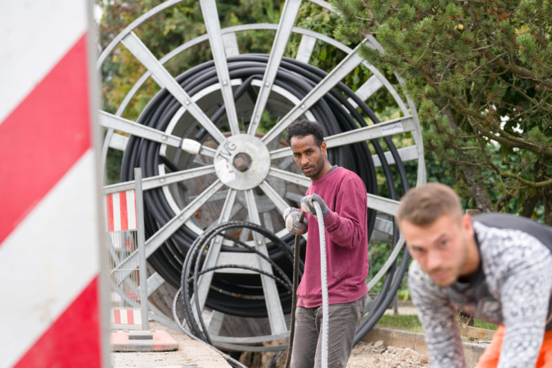 Industriefotografie: Auf einer Baustelle zur Erweiterung der Internetversorgung verlegen zwei Arbeiter ein neues Kabel. Im Hintergrund sieht man die große Trommel mit der schwarzen Erdleitung.