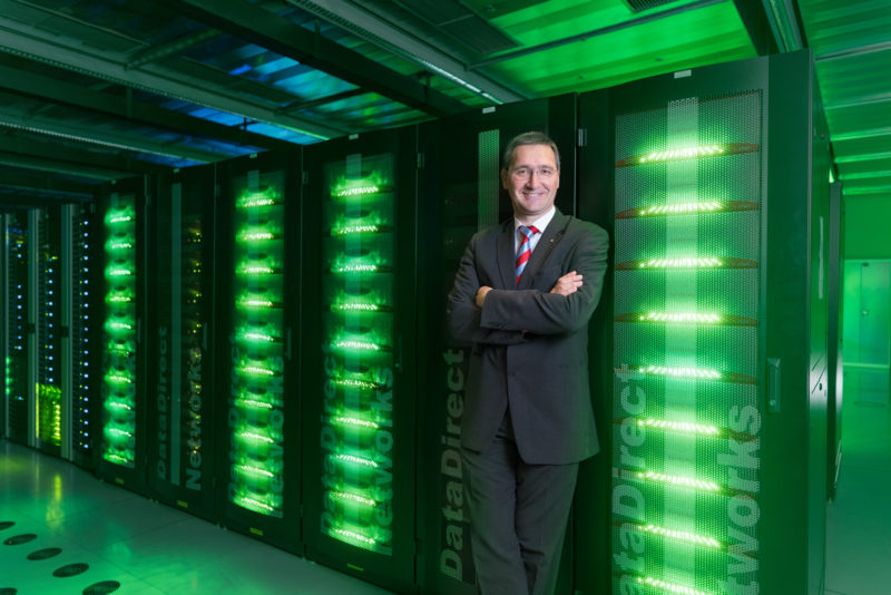 Managerportrait: A head of an IT department is leaning against one of the many green glowing black servers in his data center.