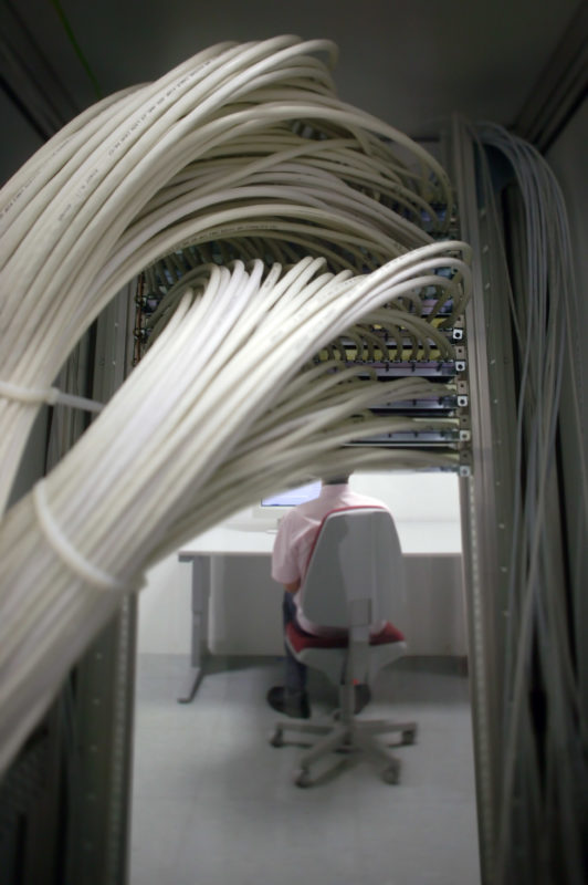 Industriefotografie: Ein Computernutzer in einem Rechenzentrum sitzt an seinem Bildschirm hinter ihm laufen dutzende Kabel in einem Rack zusammen.