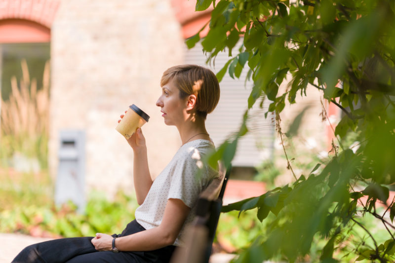Mitarbeiterfotografie: Die Mitarbeiterin eines Verlages sitzt in ihrer Pause im Grünen und trinkt einen Kaffee aus einem Pappbecher.