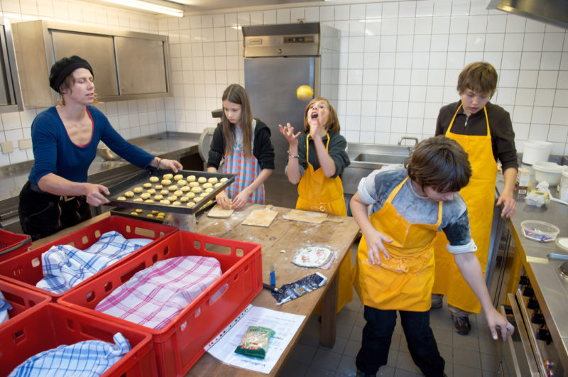 Editorial photography, subject learning and education: A school class at the cooking class under the supervision of a teacher at the Hohenlohe Open Air Museum. A student throws pastry dough in the air.