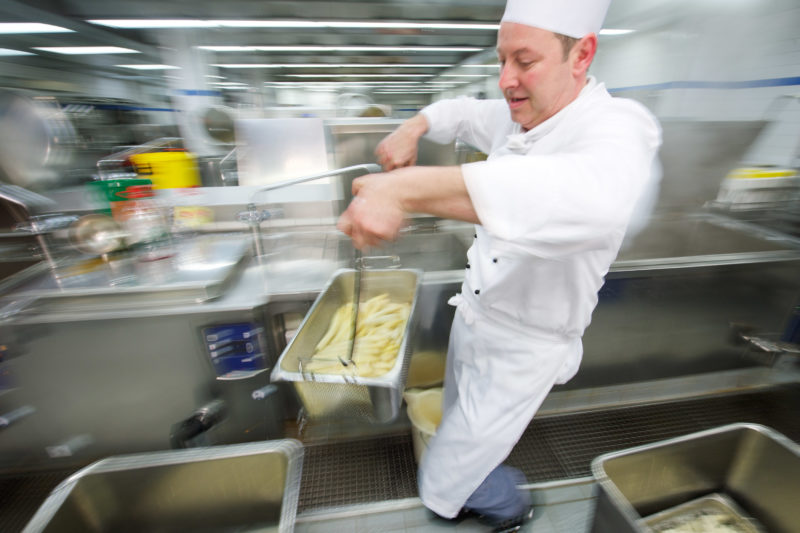 Employees photography: Employee portrait: A chef in a canteen kitchen carries the hot white asparagus that has just been cooked for further processing. The photo shows him in motion. One sees the movement through a long exposure time, which creates a blurring effect.