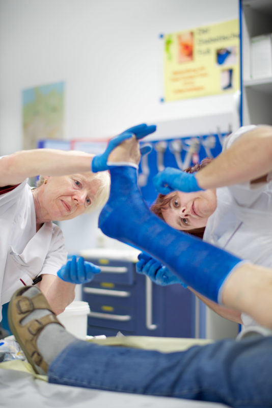 Medizinfotografie: Zwei Gesundheits- und Krankenpflegerinnen umwickeln im Gipsraum einer Klinik einen verletzten Fuß mit einem blauen Kunststoffverband.