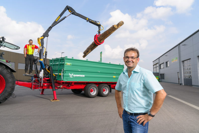 Mitarbeiterfotografie: Ein Mitarbeiter eines Betriebes für landwirtschaftliche Geräte vor einem modernen Anhänger zum Transport von Holz, welcher einen hydraulischen Kran für das Laden von Holzstämmen integriert hat.