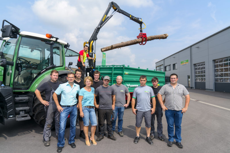 Group photo: Employees of an agricultural machinery dealer in front of one of their offered products, a trailer to transport wood on a tractor. On the crane of the hanger hangs a tree trunk.