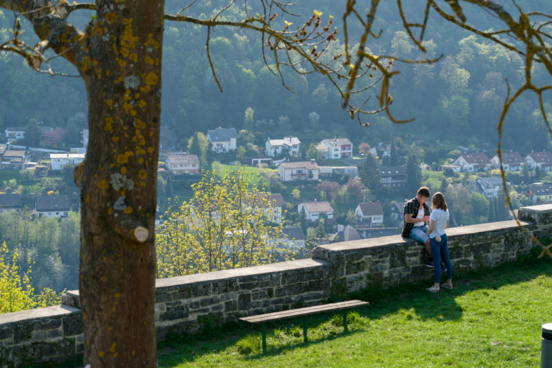 Landschaftsfotografie: Ein Teenager-Pärchen auf einem romatischen Aussichtspunkt auf einem Burggelände über einer typischen Kleinstadt in Deutschland mit viel sommerlichem Grün.