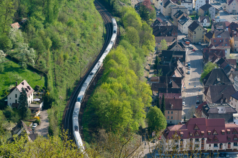 Landschaftsfotografie: Ein Intercity-Express schlängelt sich durch ein Tal. Grüne Bäume trennen die Bahnlinie von den Häusern einer Siedlung.