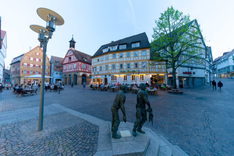 Landschaftsfotografie: Stadtansicht vom Marktplatz in Waiblingen. Das Foto wurde in der blauen Stunde aufgenommen. Vor dem Café Tagblatt sitzen viele Gäste. Im Vordergrund die Plastik Die Taubenhäusler des Künstlers Karl-Henning Seemann.