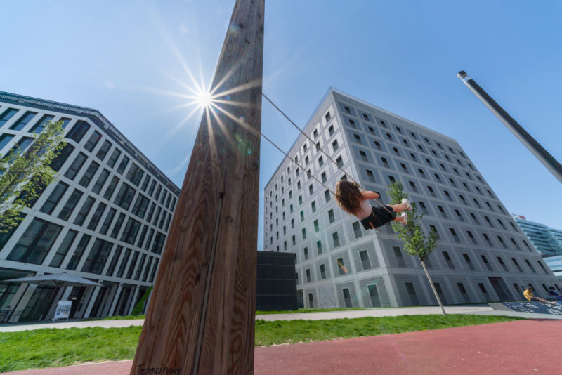 Stadtportrait Stuttgart: Landschaftsfotografie: Vor den modernen Gebäuden am Pariser Platz in Stuttgart schaukelt ein Kind in der Sonne.