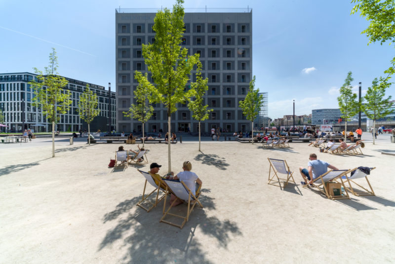 Landscape photogaphy:  On the sunlit Pariser Platz in Stuttgart, people sit in beach chairs. In the background, the striking building of the municipal library rises into the blue sky. Green young trees line the scene.