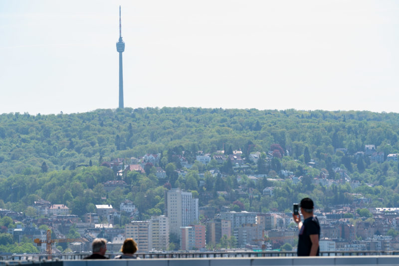 Stadtportrait Stuttgart: Landschaftsfotografie: Ein Blick über die Stadt Stuttgart. Man sieht die umliegenden bebauten aber grünen Hänge und darüber den Fernsehturm, eines der Wahrzeichen der Stadt. Im Vordergrund fotografiert ein Tourist ein Selfie.