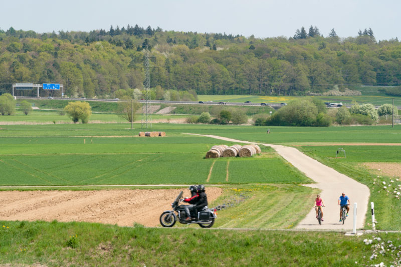 Landschaftsfotografie: Eine typische Wald-, Wiesen- und Felderlandschaft in Süddeutschland. Dazwischen Radfahrer und ein Motorrad auf Ausflug. Dahinter eine Autobahn. Große Strohrollen liegen in einem grünen Feld.
