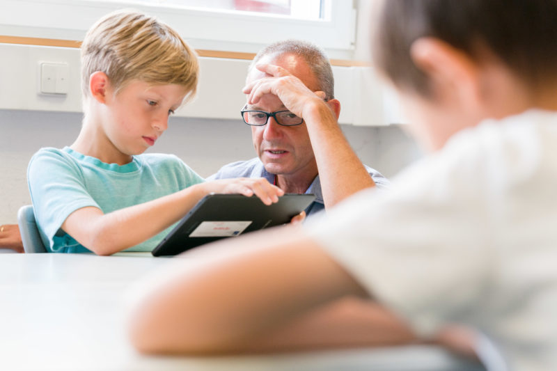 Reportagefotografie: Ein Lehrer bei der Arbeit: Der Lehrer hilft einem Schüler bei der Arbeit mit einem Tablet Computer. Er stützt dabei seinen Kopf nachdenklich auf.