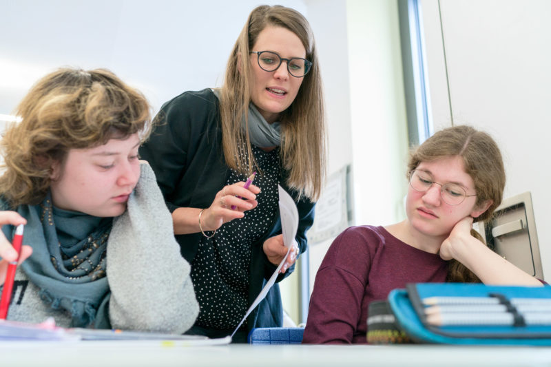 Reportagefotografie zum Thema Lernen und Bildung: Während des Unterrichts schaut eine Lehrerin an einer Berufsschule zwei Schülerinnen über die Schulter und hilft ihnen.