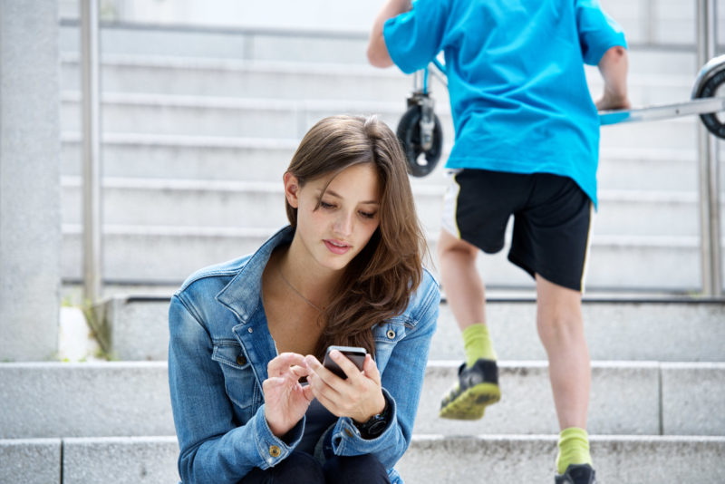 Lifestyle-fotografie: Eine junge Frau sitzt draußen auf einer Treppe und schaut auf ihr Smartphone während im Hintergrund ein Junge die Treppe hoch rennt.