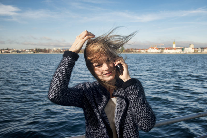 Lifestylefotografie: Eine Frau mit wehenden Haaren telefoniert mit dem Smartphone an Bord einer Fähre auf dem Bodensee. Ihre Haare wehen so sehr im Wind, dass man ihr Gesicht kaum sieht.