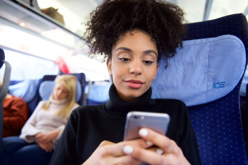 Lifestyle photography: A woman sits in a ICE train and types into her smartphone. In the background a second woman is sleeping.