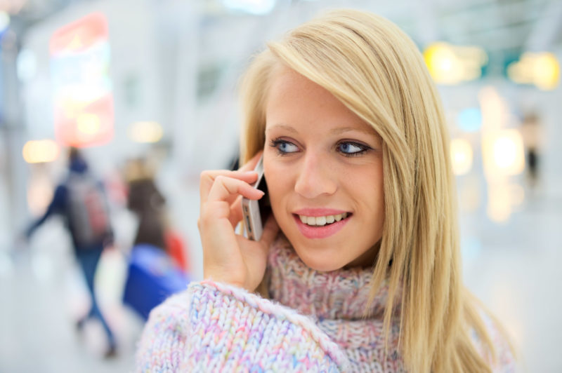 Lifestyle photography: At an airport a young blonde woman speaks into her cell phone. In the background travelers with suitcases pass by.