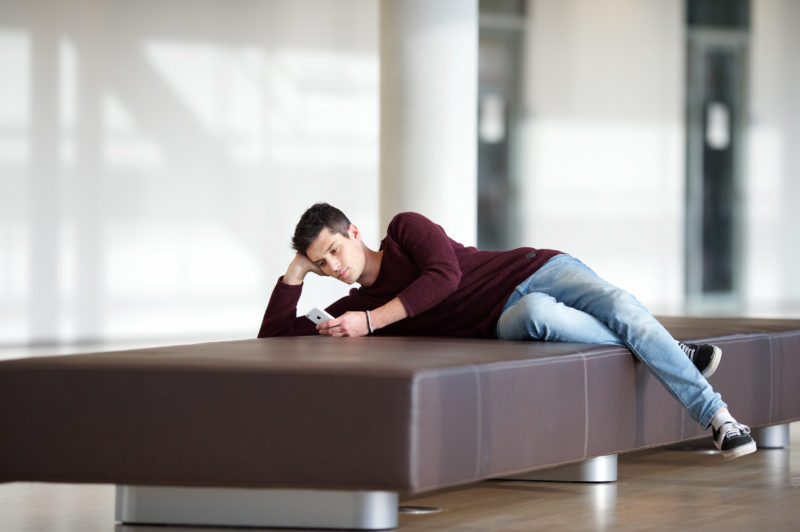 Lifestyle-Fotografie: In einem öffentlichen Gebäude liegt ein junger Mann bequem auf einer Bank und schaut in sein Smartphone.